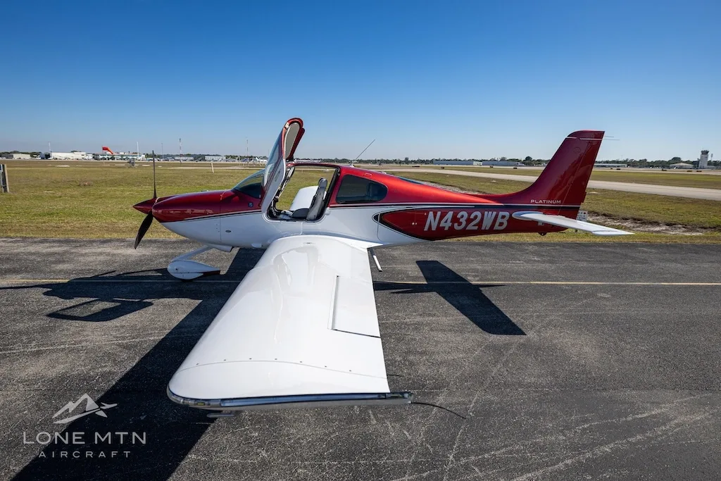 An overhead front-side view of the red and white Cirrus SR20 aircraft (N432WB) with its doors open, parked on an airport tarmac.