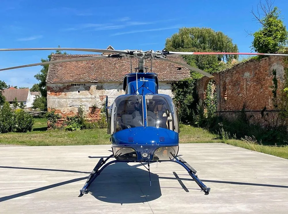 A blue Bell 505 helicopter is shown from the front, parked on a concrete pad with an old building in the background.
