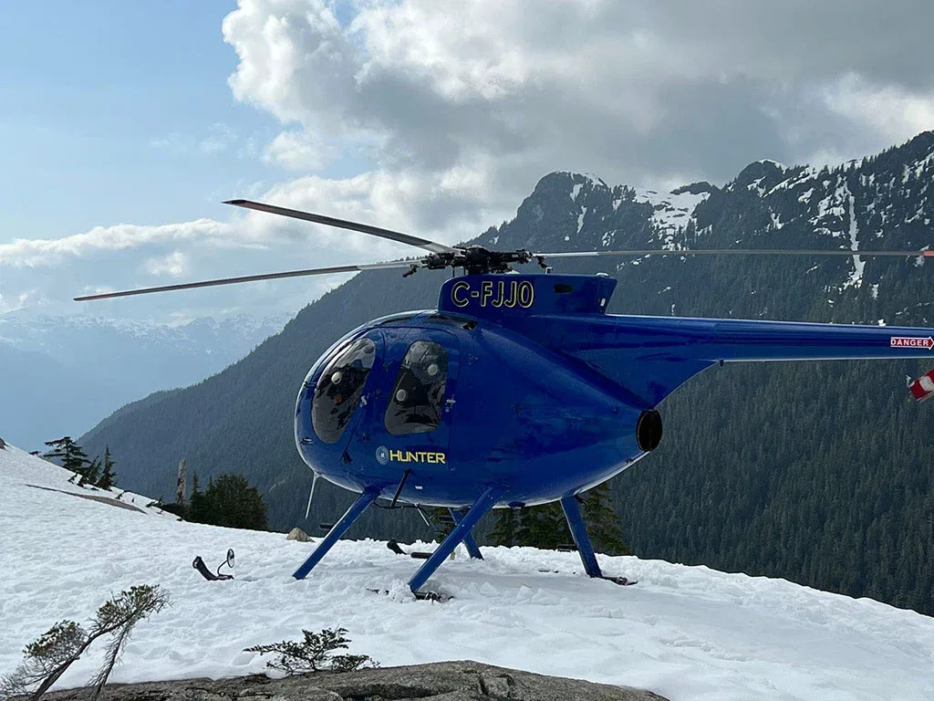 A blue helicopter with registration C-FJJ0 is parked on a snow-covered mountain slope with a cloudy sky.