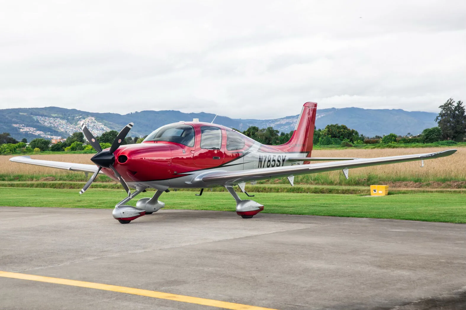 A red and silver Cirrus SR22T aircraft, registration N785SY, parked on an airfield with mountains in the background.