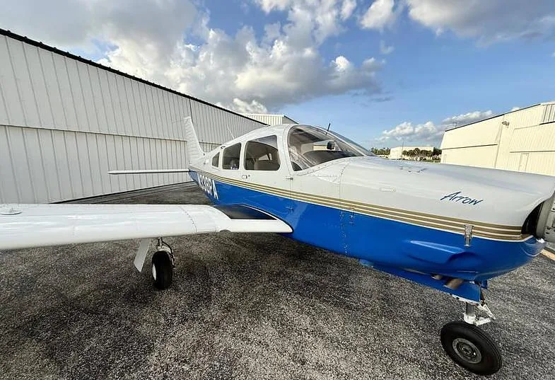 A close-up, front-right view of a white and blue Piper Arrow aircraft with registration N338PA parked on an asphalt tarmac.