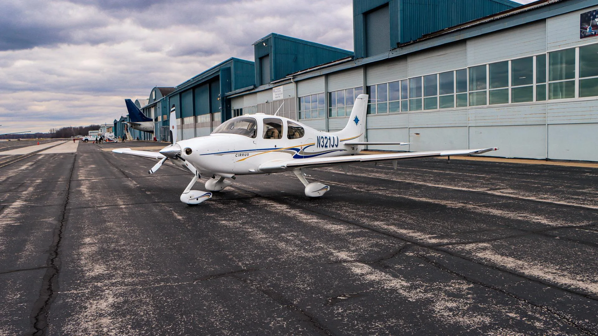 A white Cirrus aircraft, registration N321JJ, with blue and gold stripes, is parked on an airport tarmac.