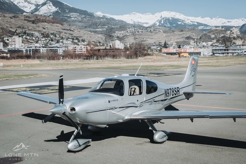 A silver Cirrus SR22 aircraft (N978SR) parked on an airfield with snow-capped mountains and a town in the background.