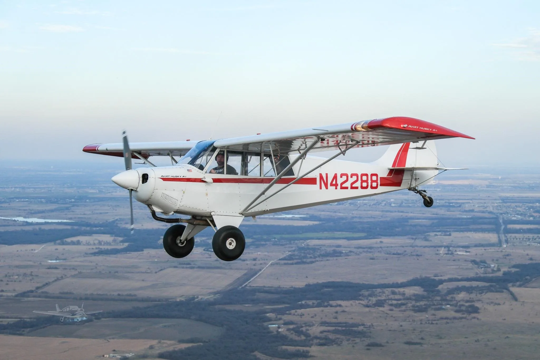 A white and red Aviat Husky A-1 aircraft with registration N42288 flies over a rural landscape, with another aircraft faintly visible below.