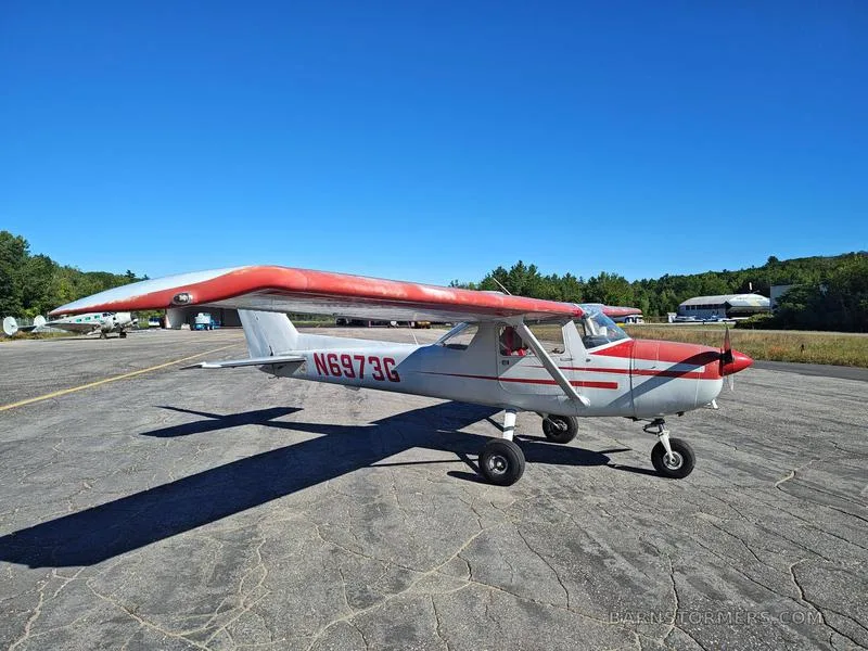 A white and red Cessna 150/152 aircraft with registration N6973G is parked on an asphalt tarmac under a clear blue sky.