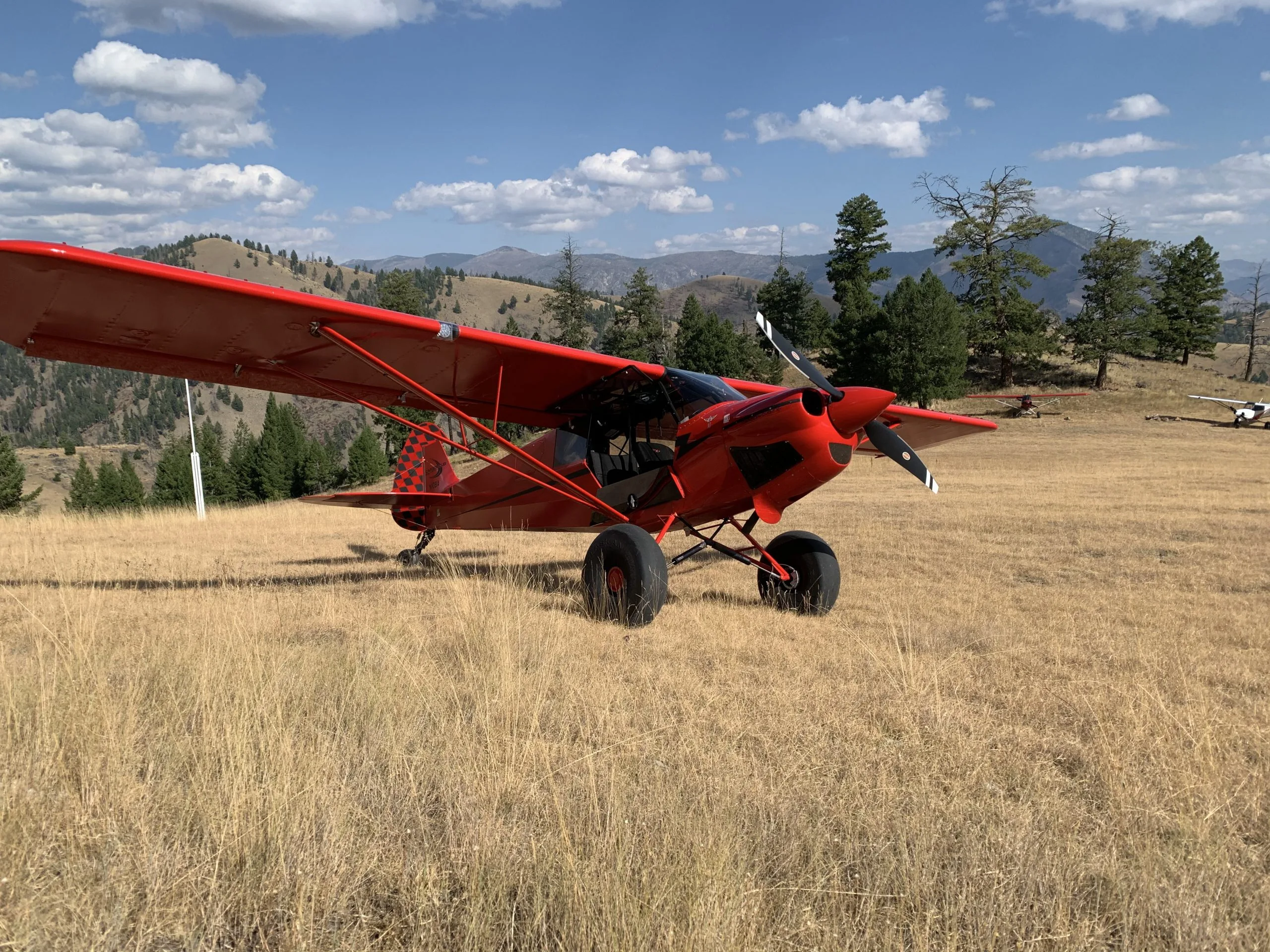 A red bush plane with large tundra tires and a checkered tail is parked on a grassy mountain airstrip under a blue sky.