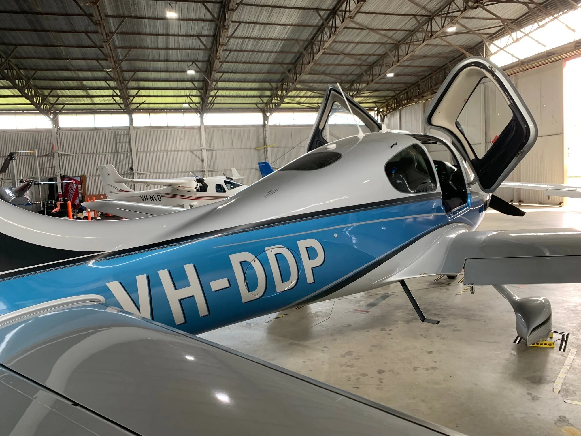 A silver and blue Cirrus SR22T aircraft with open doors, registration VH-DDP, parked inside a hangar.