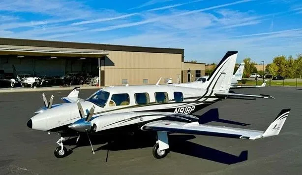 A white and black twin-engine Piper aircraft, N91BP, is parked on an airfield with a hangar in the background.