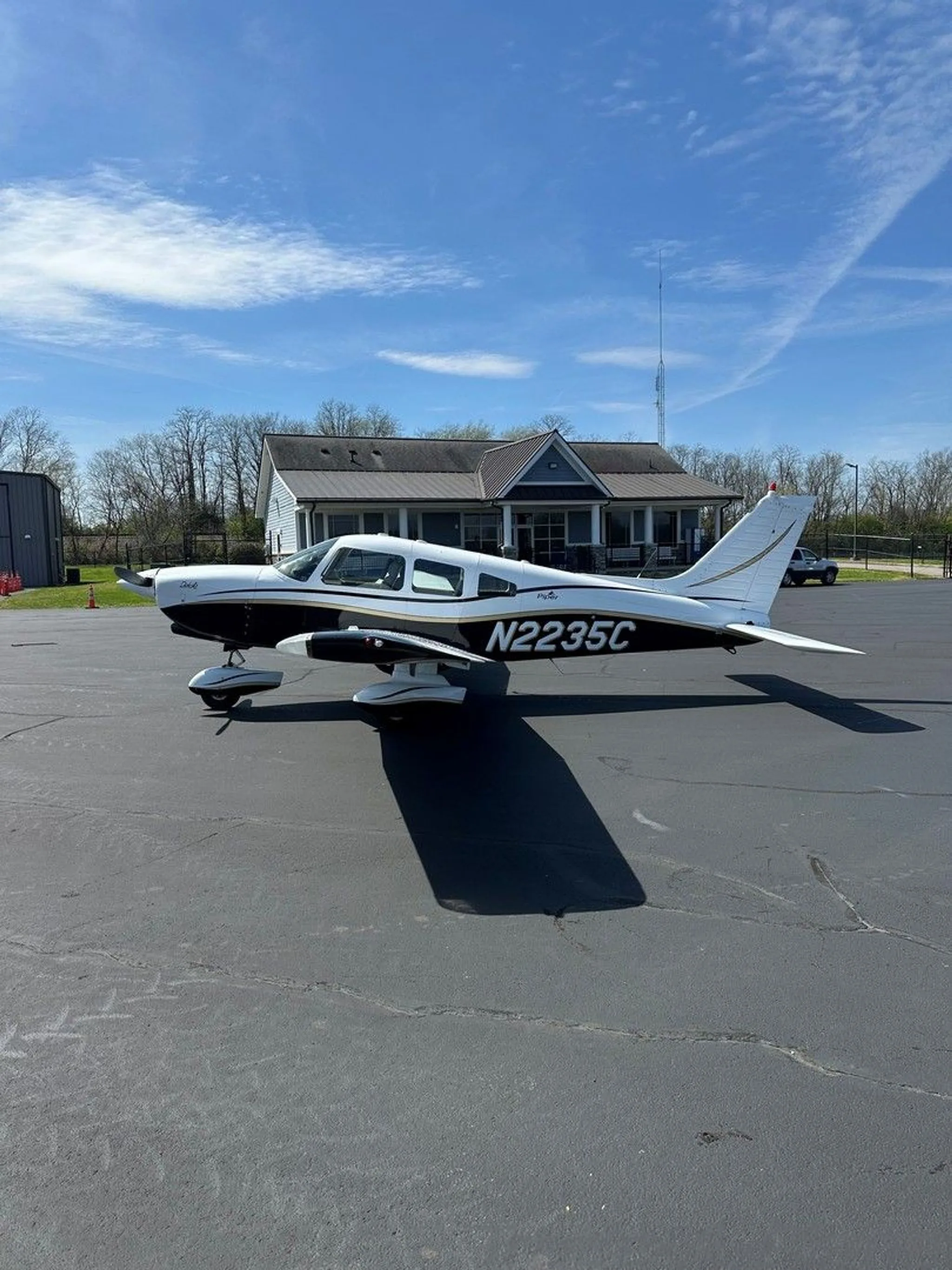 A white and black Piper aircraft with gold trim, registration N2235C, parked on an asphalt ramp with a building in the background.