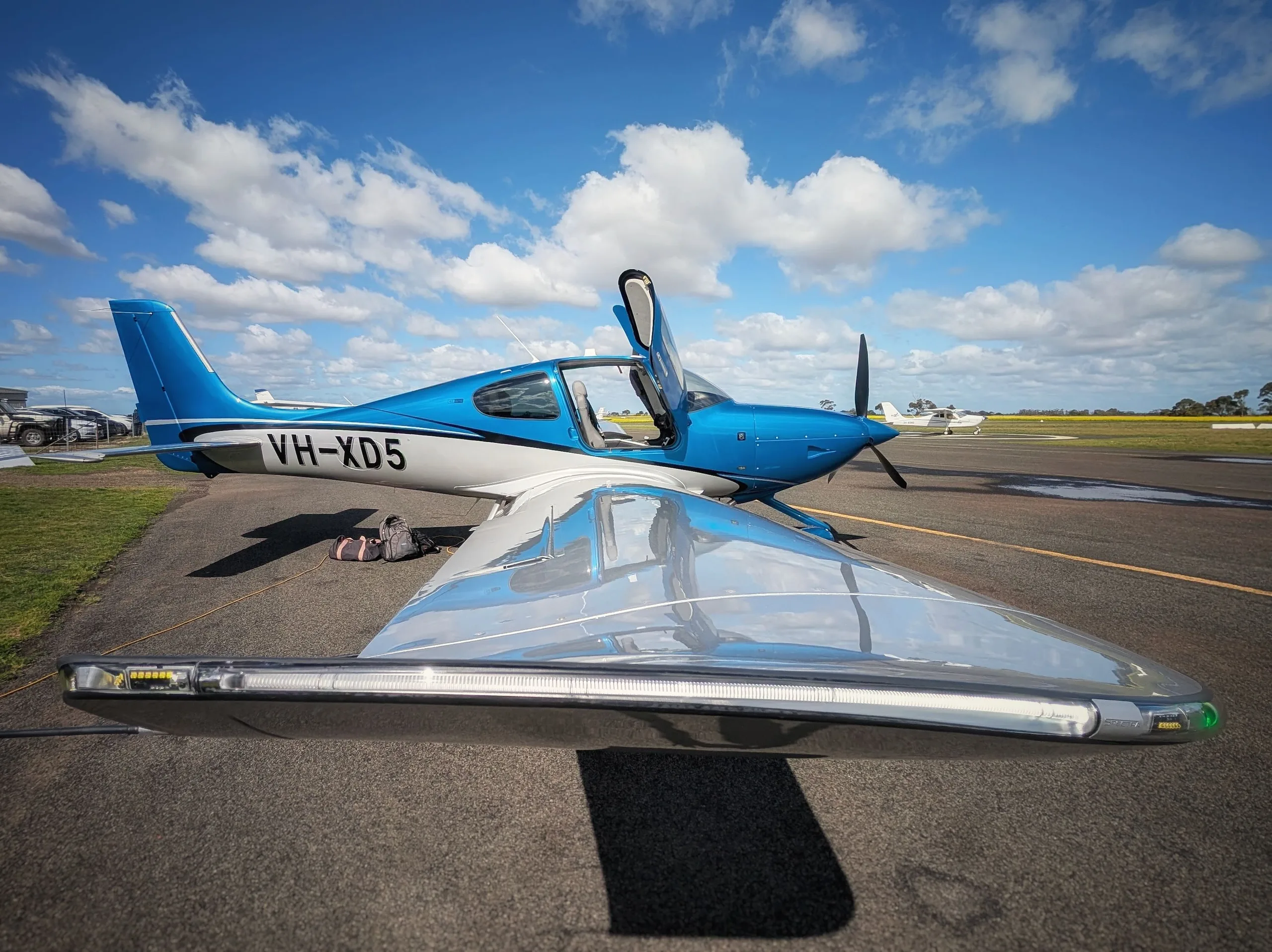 A blue and white single-engine Cirrus SR22 aircraft with registration VH-XD5 parked on an airfield with its doors open.