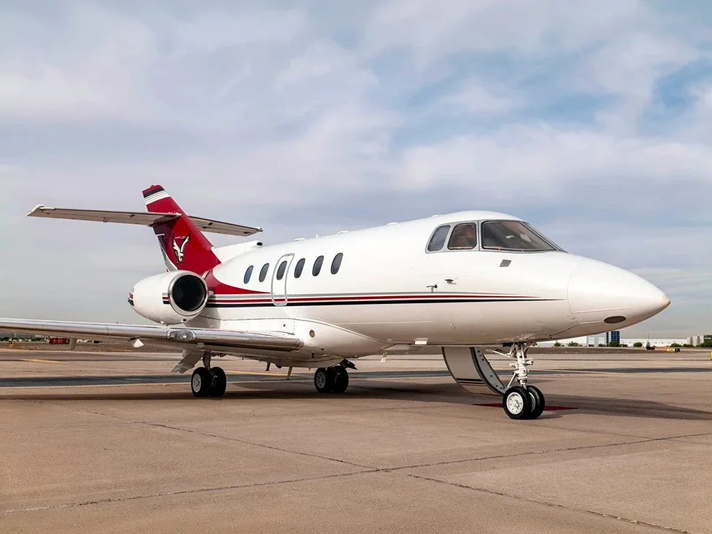 A white business jet with a red tail featuring a bull logo and red and black stripes, parked on an airport tarmac.