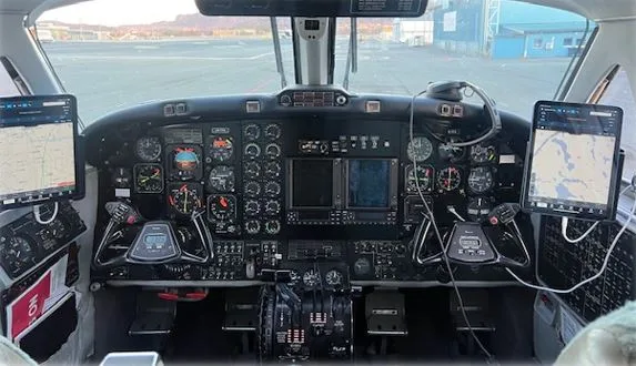A wide view of an aircraft cockpit with a complex instrument panel, two yokes, and two tablets mounted on either side, looking out onto an airport tarmac.