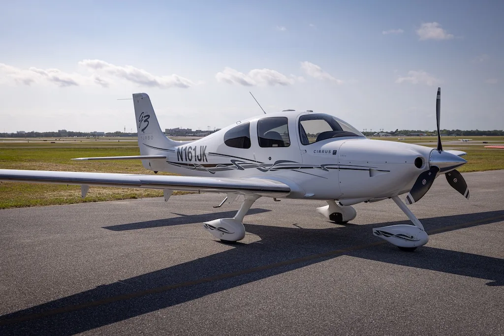 A white Cirrus SR22 Turbo aircraft with black and grey stripes, registration N161JK, parked on an asphalt tarmac under a blue sky.