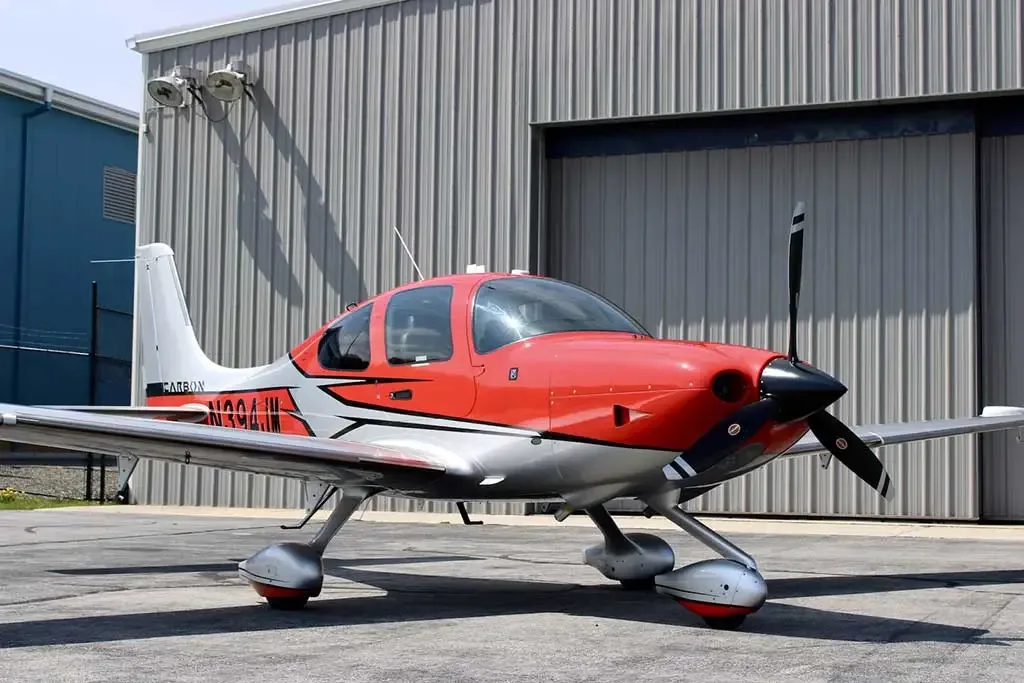A red and silver Cirrus SR22 aircraft with registration N394JM parked in front of a hangar.