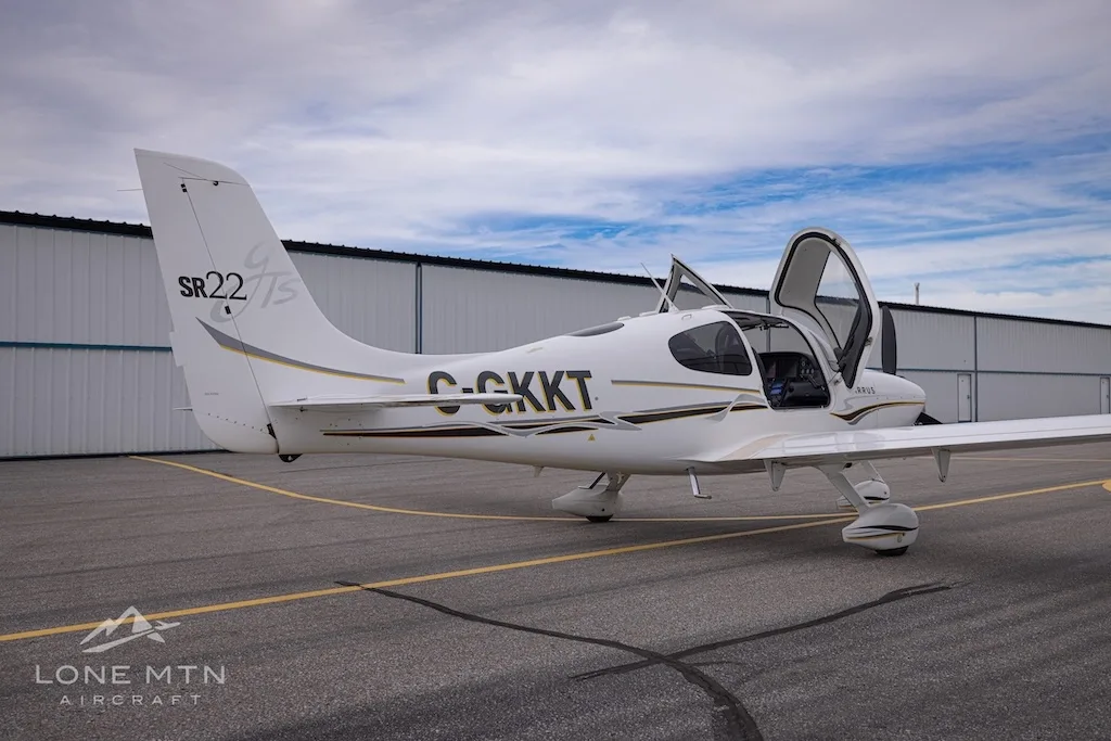 A rear three-quarter view of a white Cirrus SR22 aircraft with registration C-GKKT, doors open, on an airfield.