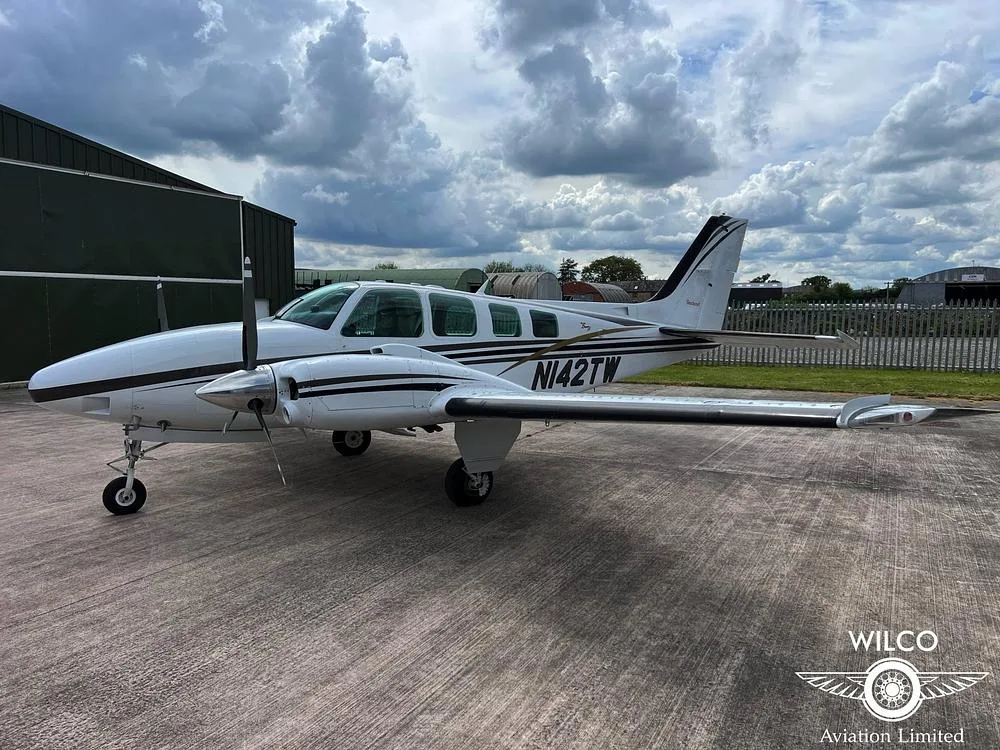 A white Beechcraft Baron N142TW with black and gold stripes parked on a tarmac, viewed from the left side.