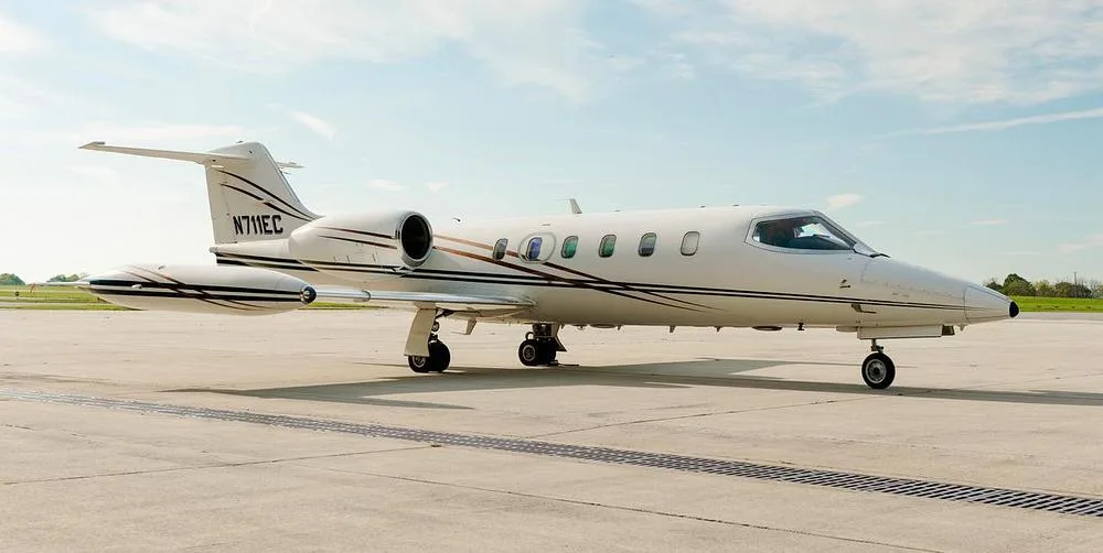 A white Learjet 35A, registration N711EC, with brown and black stripes, parked on an airfield under a partly cloudy sky.
