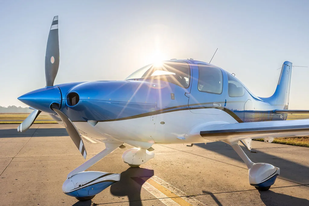 A blue and white Cirrus SR22 aircraft parked on an airfield with the sun flaring over its nose.