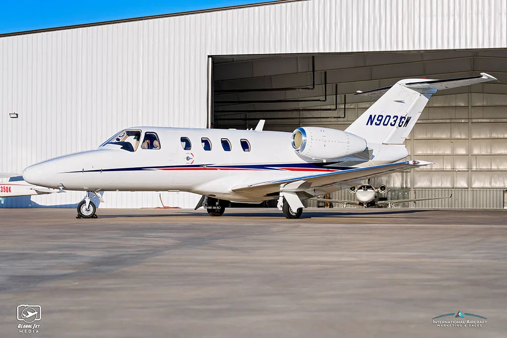 A side view of a white Cessna Citation CJ3 (N903GW) with blue and red stripes parked on an tarmac.