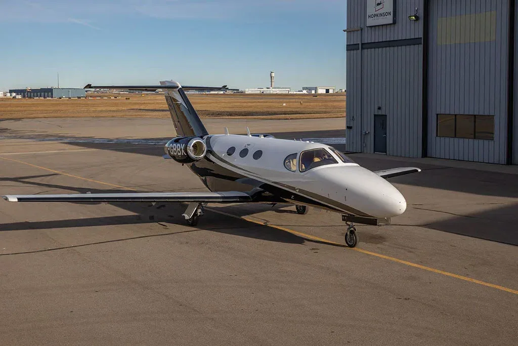 A white and black private jet, registration C-GBBK, parked on an asphalt apron with a hangar and airport buildings in the background.