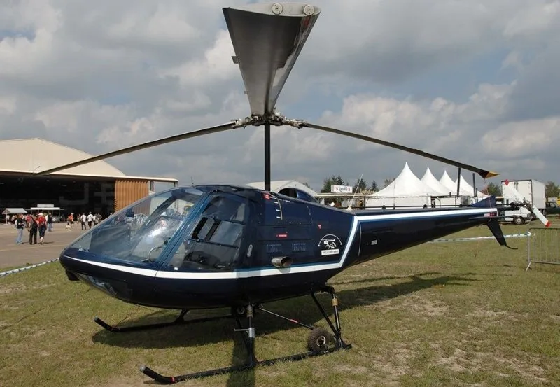 A dark blue helicopter with a white stripe is parked on a grassy field at an airfield with hangars and tents in the background.