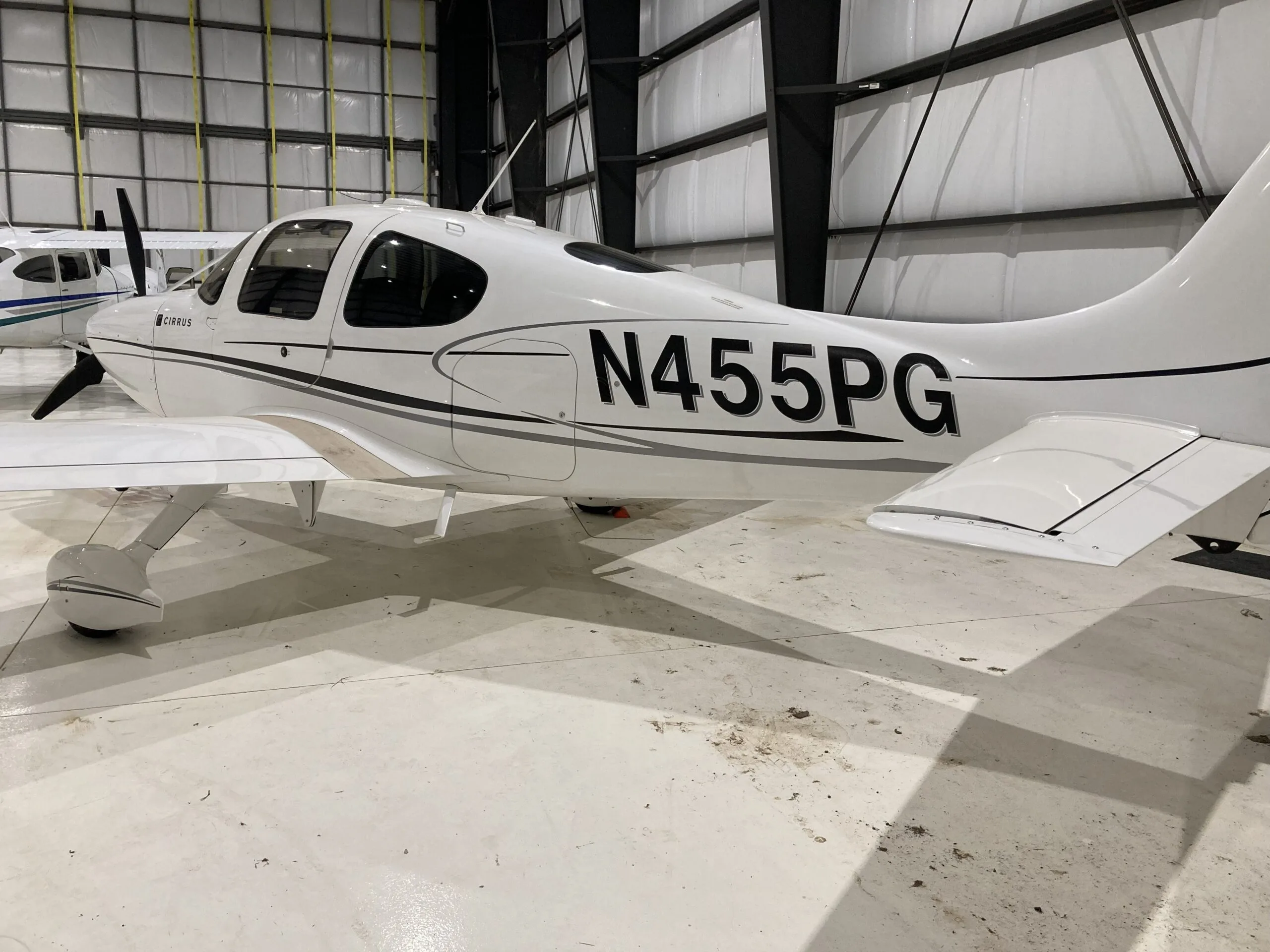 Rear-side view of a white Cirrus SR22 aircraft with black and grey stripes, registration N455PG, in a hangar.