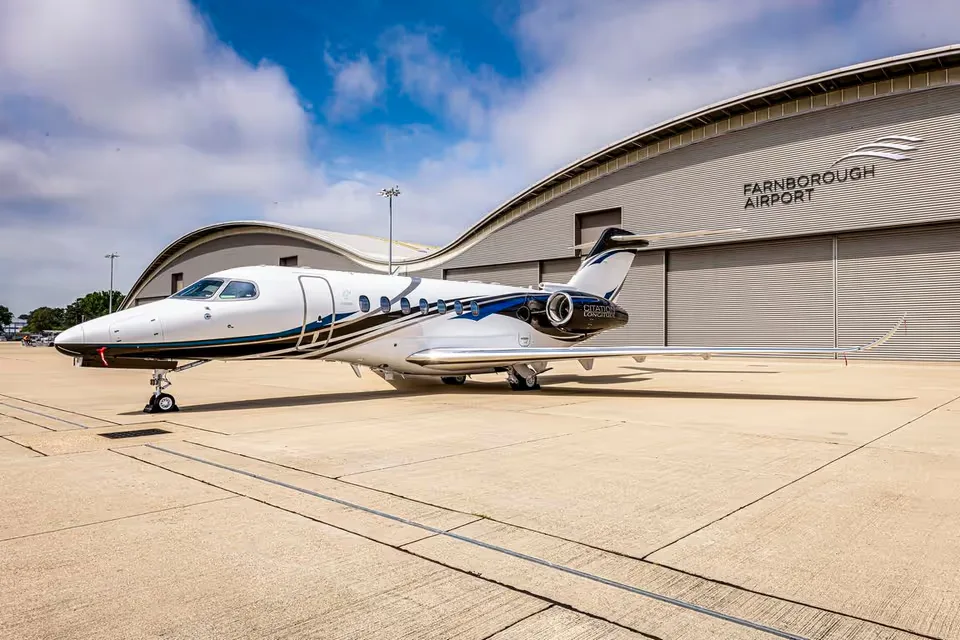 A white Cessna Citation Longitude with blue and gold stripes is parked on an apron in front of a Farnborough Airport hangar.