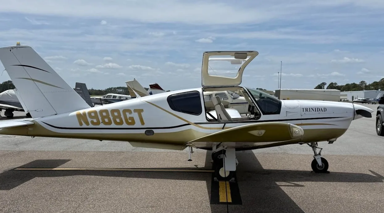 A side view of the white and gold Socata TB-20 Trinidad aircraft with its doors open on an airfield.