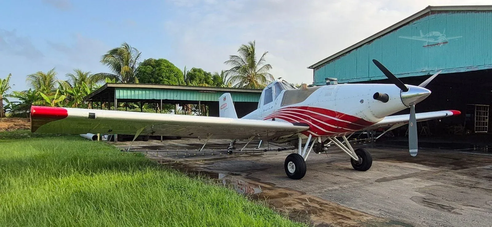 A white agricultural aircraft with red stripes and red wingtips is parked on a concrete pad next to a green hangar with palm trees in the background.