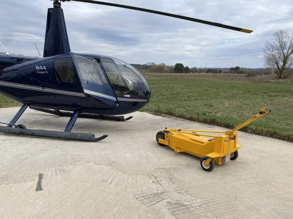 The front-left side of a dark blue Robinson R44 Clipper II helicopter, N144LR, is shown on a concrete pad next to a yellow ground handling cart.
