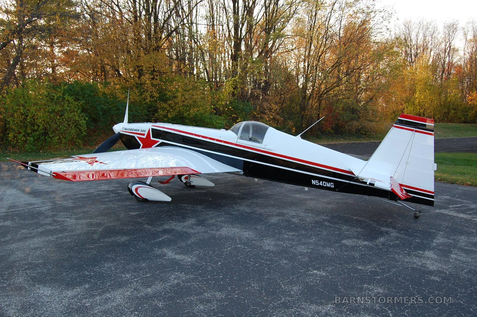 A white, black, and red Staudacher 300 aerobatic aircraft with tail number N54DMG is parked on an asphalt surface with autumn trees in the background, viewed from the side.