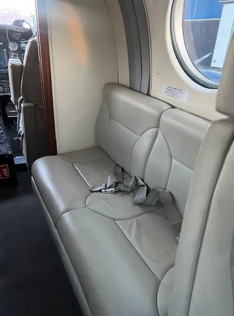 A view of the light grey leather bench seat in the aircraft cabin, with a window and part of the cockpit visible in the background.