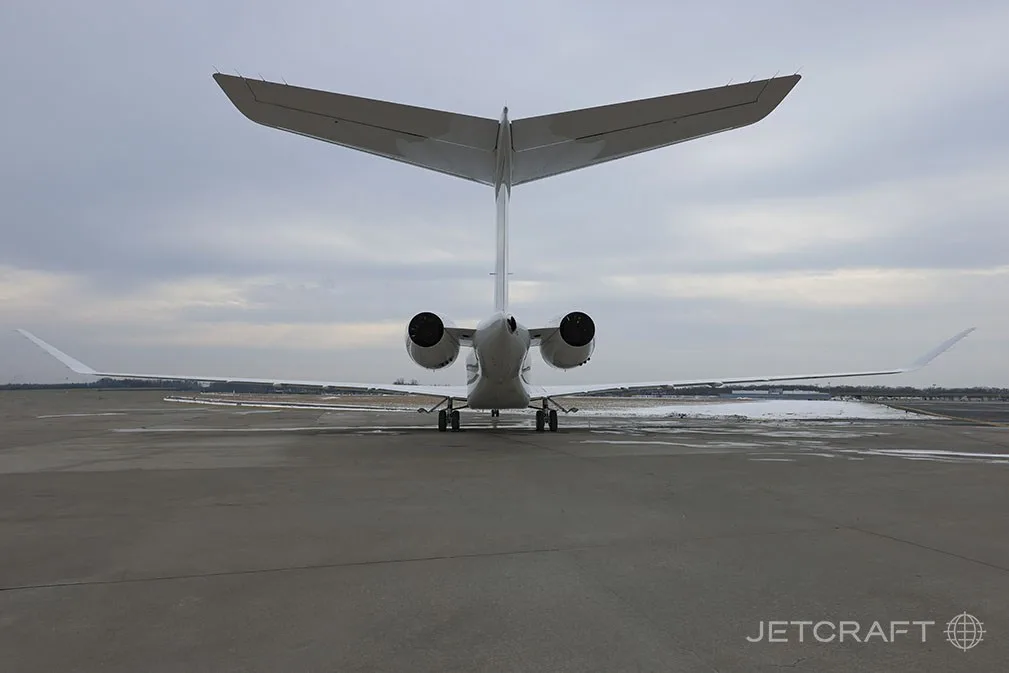 The rear view of a white Bombardier Global 7500 business jet parked on a tarmac with snow in the distance.