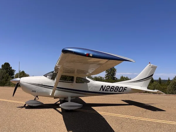A white Cessna 182Q Skylane with a blue wing and grey stripes, parked on an asphalt surface under a clear blue sky.