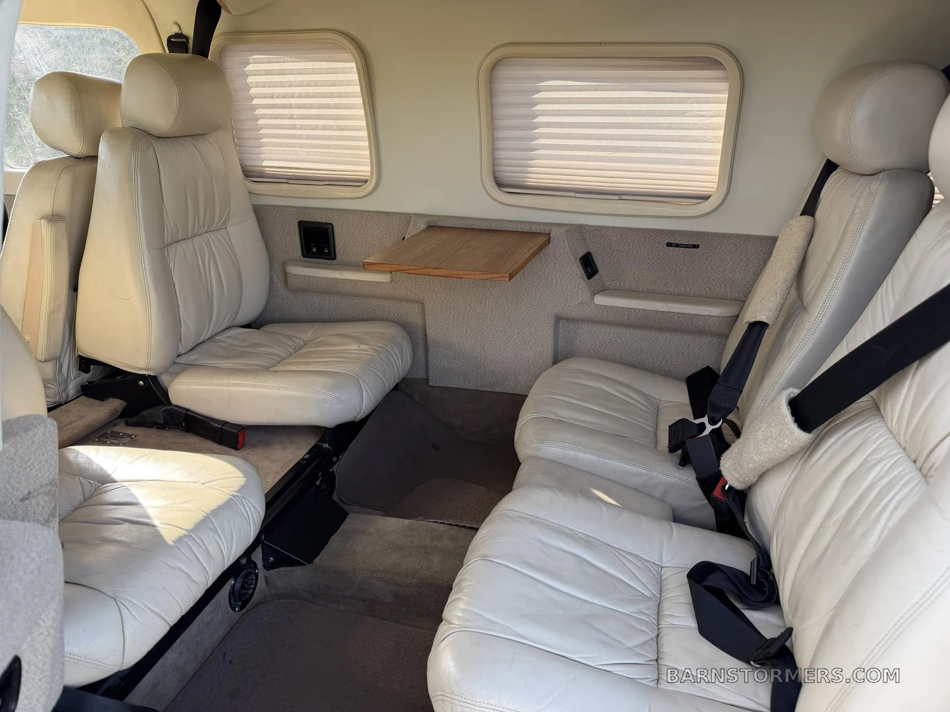 The rear cabin interior of a Piper Saratoga II TC, showing light-colored leather seats, window shades, and a small fold-out table.