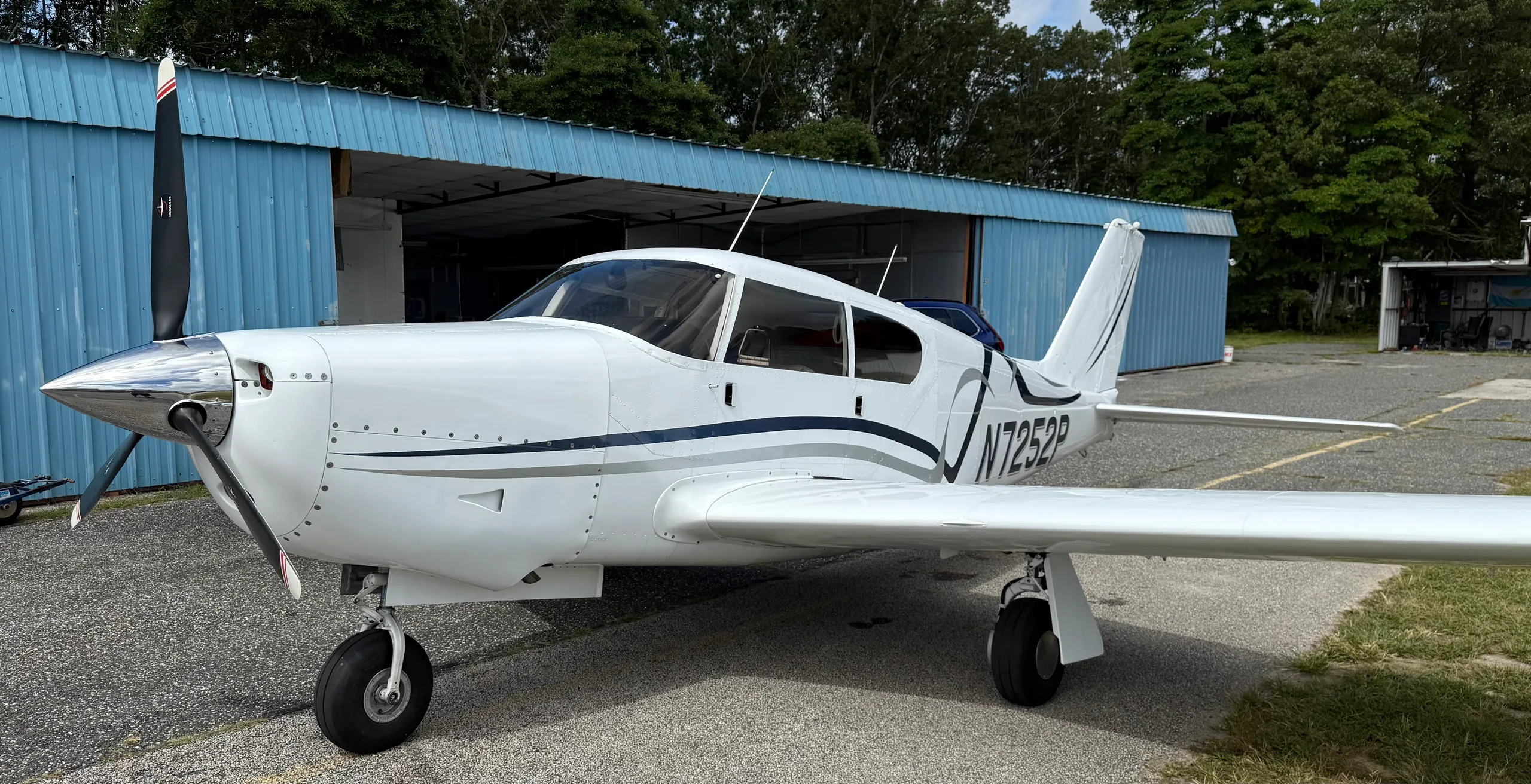 Front-left view of a white single-engine aircraft with blue and gray stripes, registration N7252P, parked on an asphalt ramp in front of a blue hangar.
