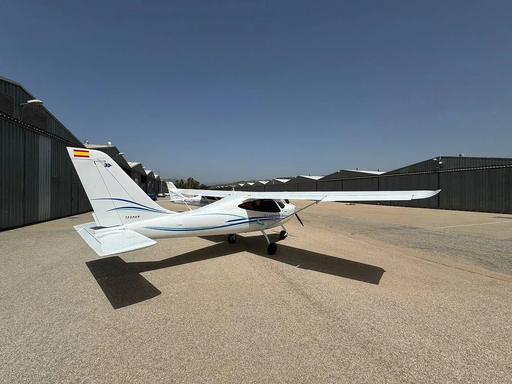 A rear-quarter view of a white Tecnam P2008 aircraft with blue stripes and a Spanish flag on the tail, parked on an airfield.