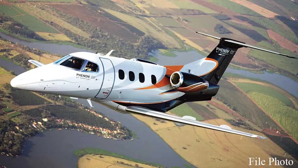 An Embraer Phenom 100 business jet with a white, orange, and black paint scheme flies over a landscape of fields and rivers.
