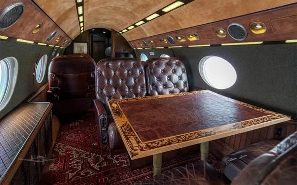 A view of the aircraft cabin with a large wooden table, brown leather seats, and a patterned red rug.