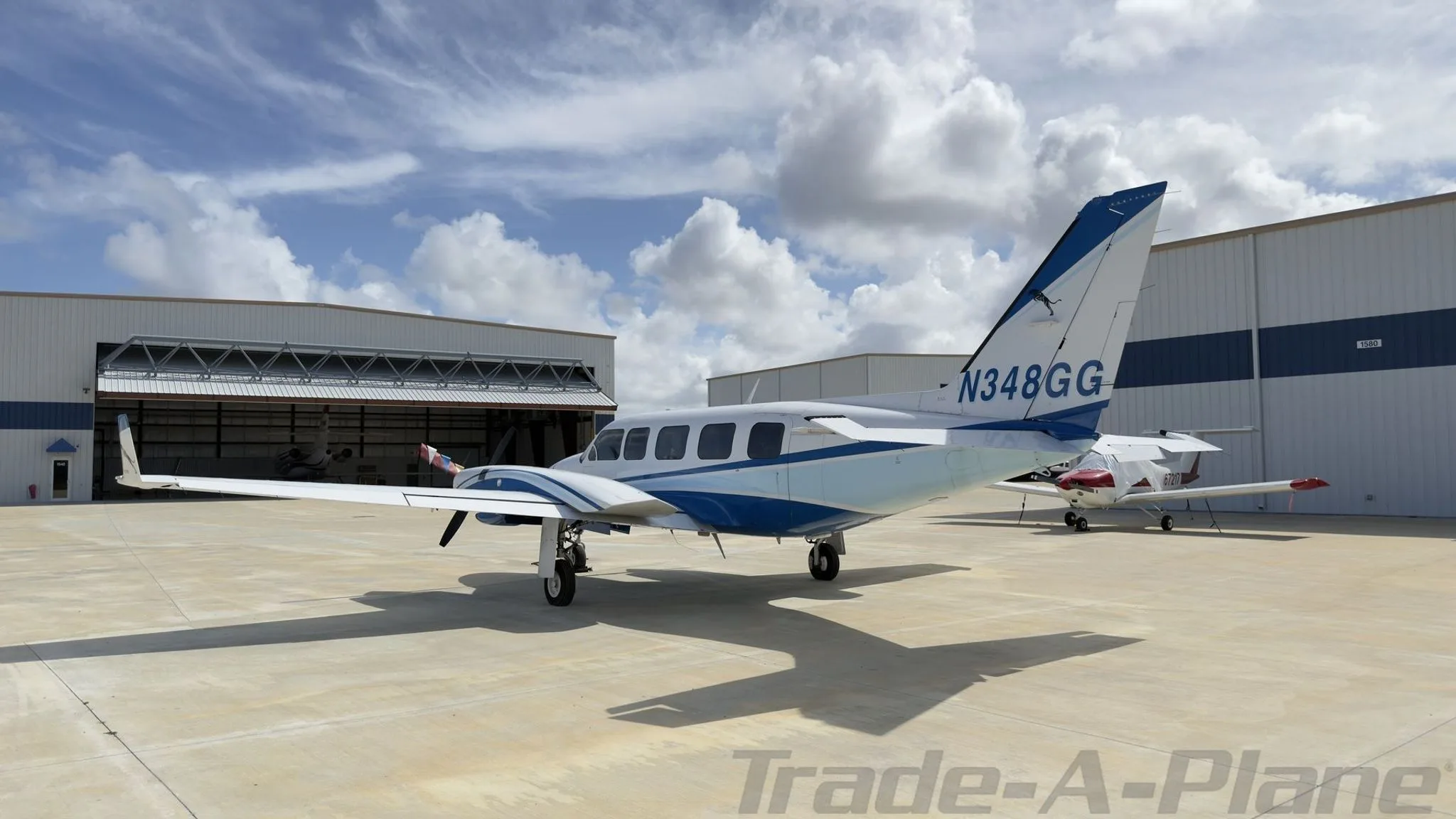 Rear-side view of a white and blue twin-engine propeller aircraft, N348GG, parked on a concrete ramp with hangars in the background.