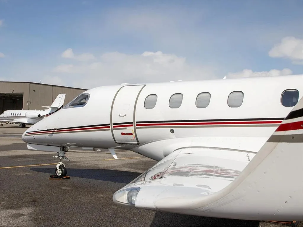 A close-up side view of a white Embraer Phenom 300 business jet with red and black stripes, parked on an airfield with a hangar in the background.