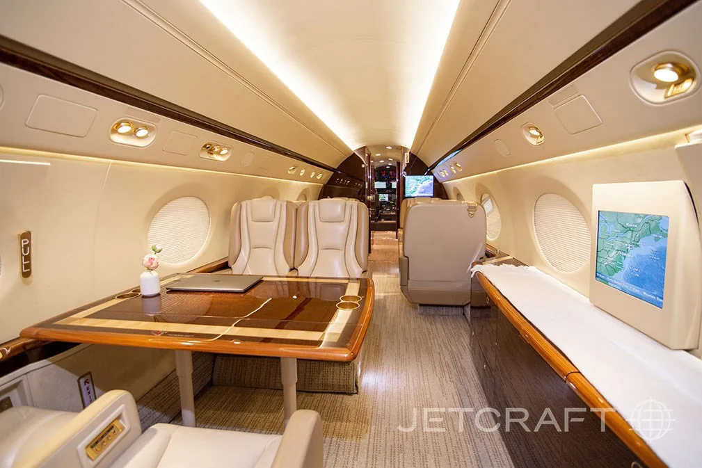 The spacious interior of a business jet cabin featuring a large wooden table and comfortable seating.