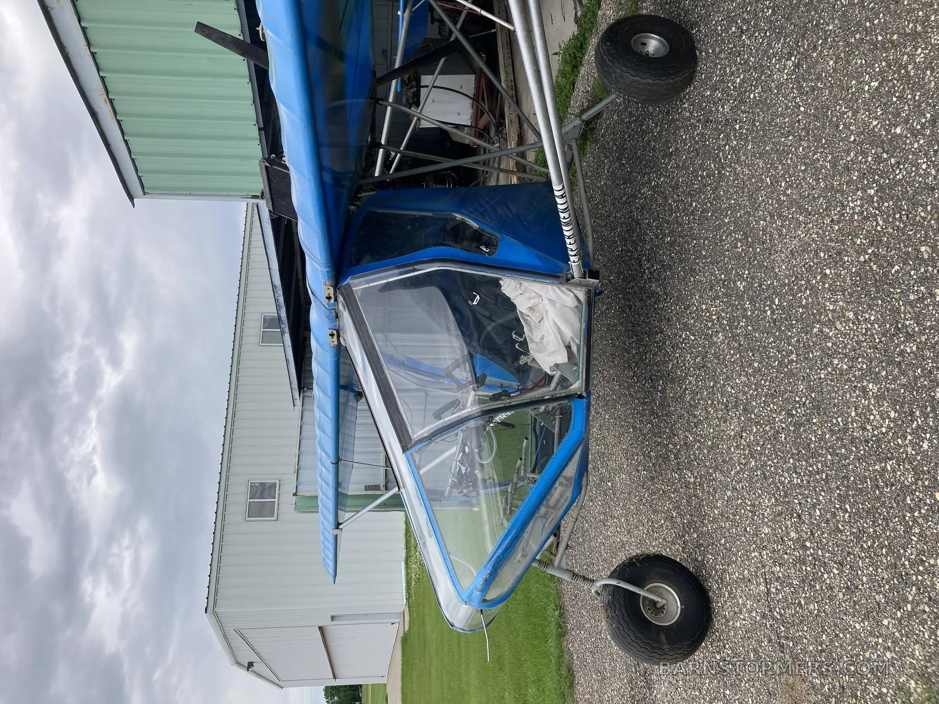 A blue ultralight aircraft with an open cockpit, parked on asphalt next to a hangar.