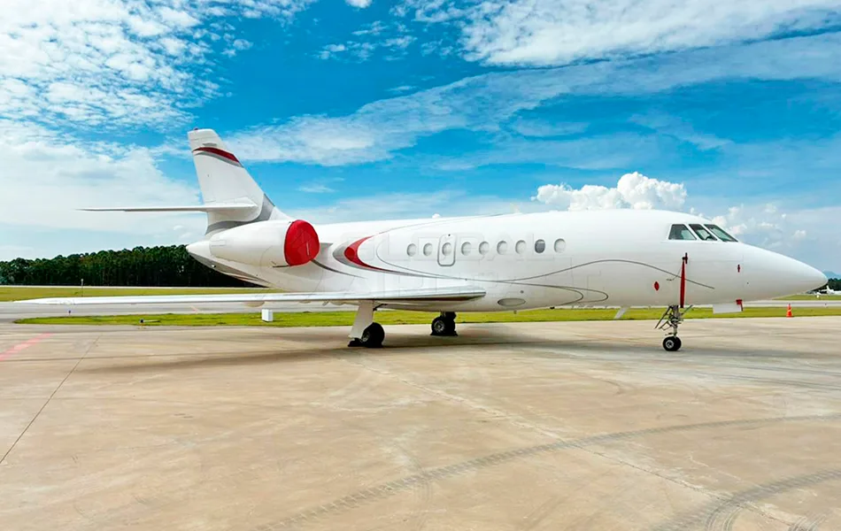 A white Dassault Falcon business jet with red engine covers and a subtle grey/red stripe parked on an airport tarmac under a blue sky.