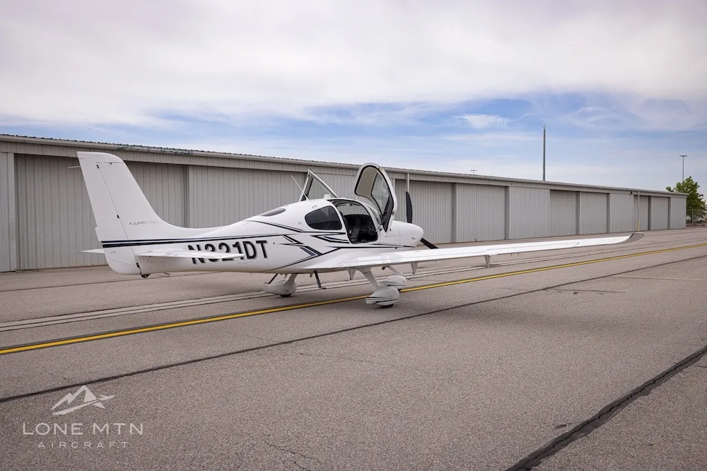 Rear-quarter view of a white Cirrus SR22 aircraft with black and grey stripes, doors open, parked on an asphalt ramp.