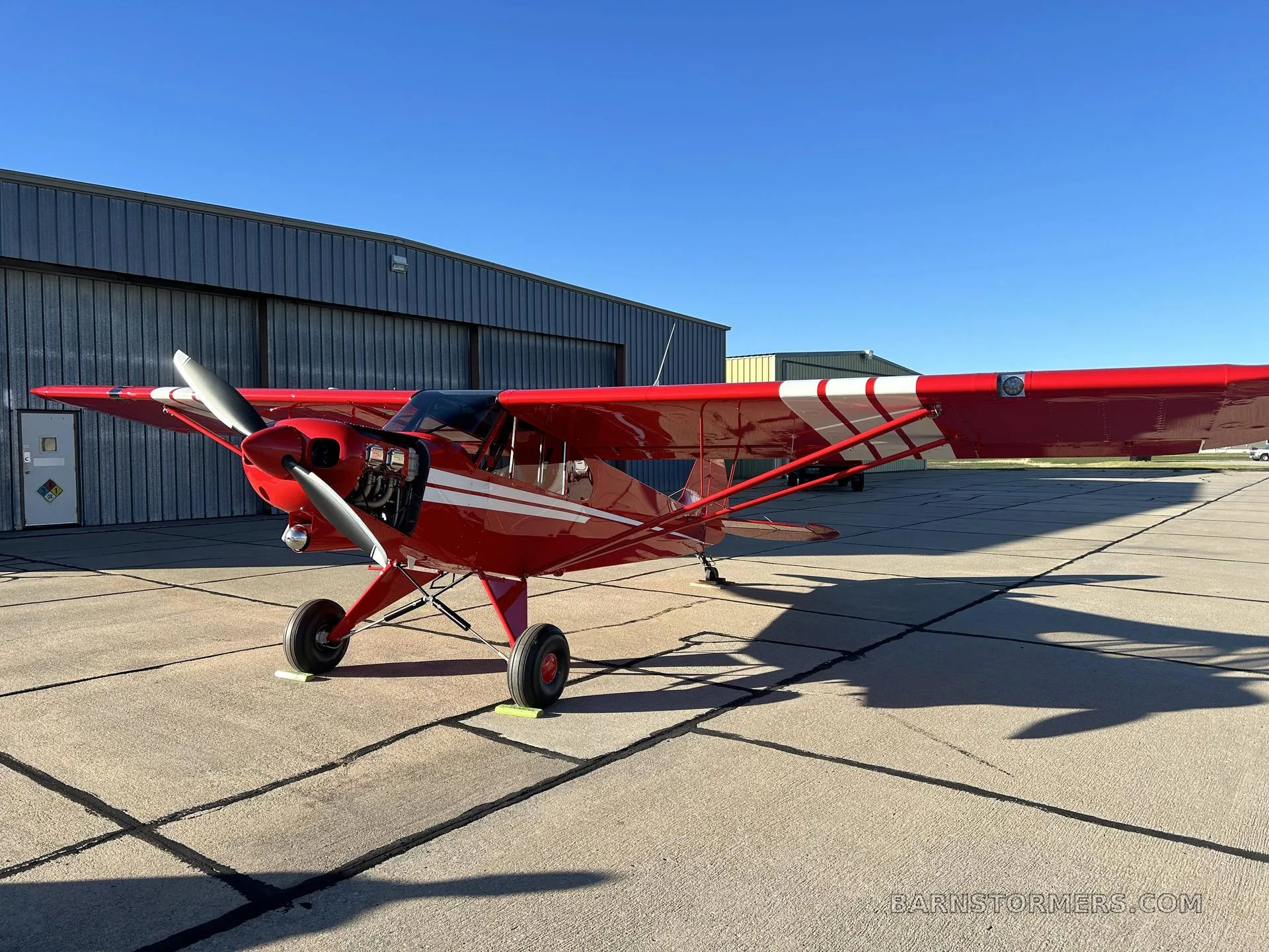 A red PA-18 Supercub on the tarmac in front of a hangar.