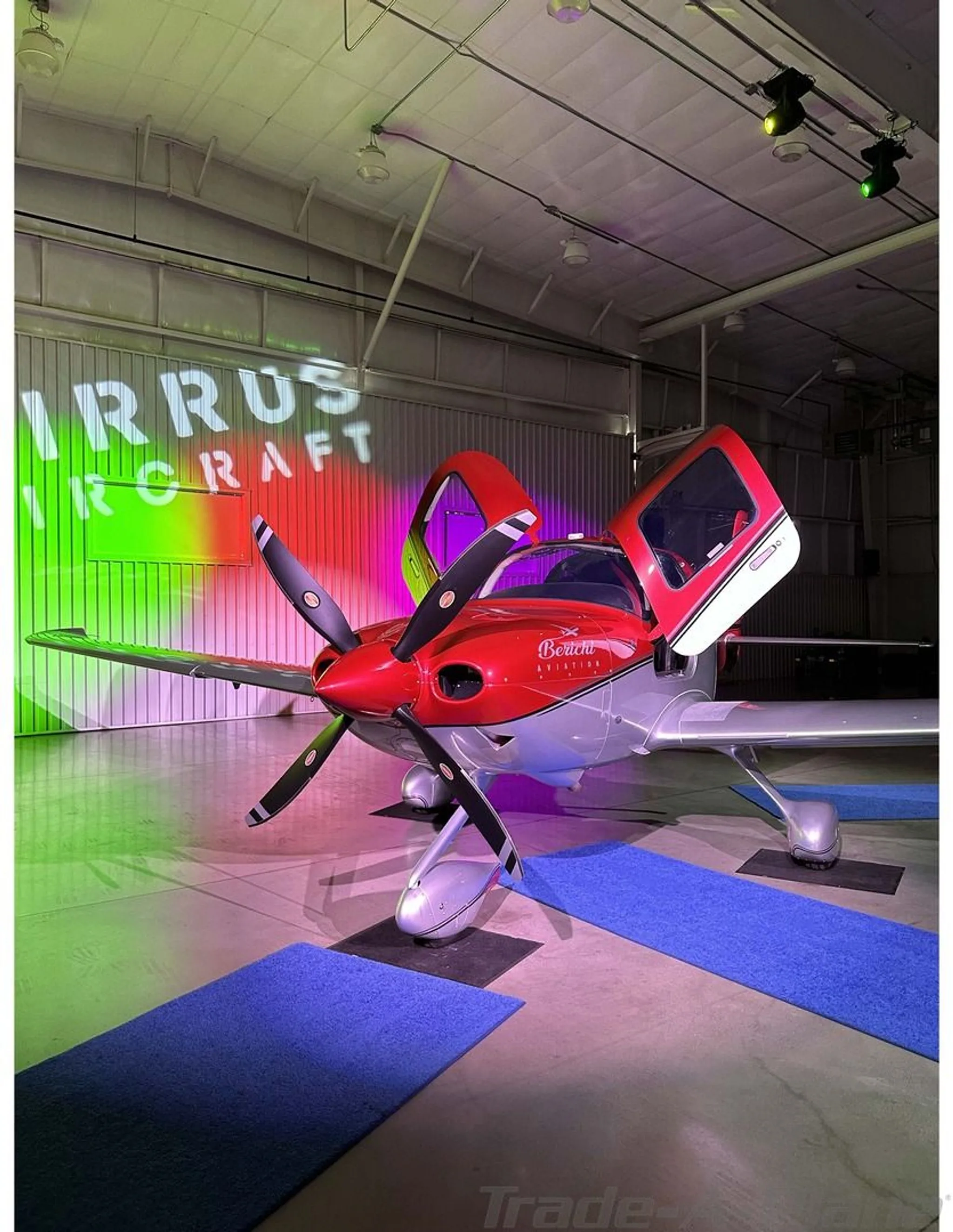 A front-quarter view of the red and silver Cirrus SR22 aircraft, N657CB, with its doors open, in a hangar with colorful lighting.