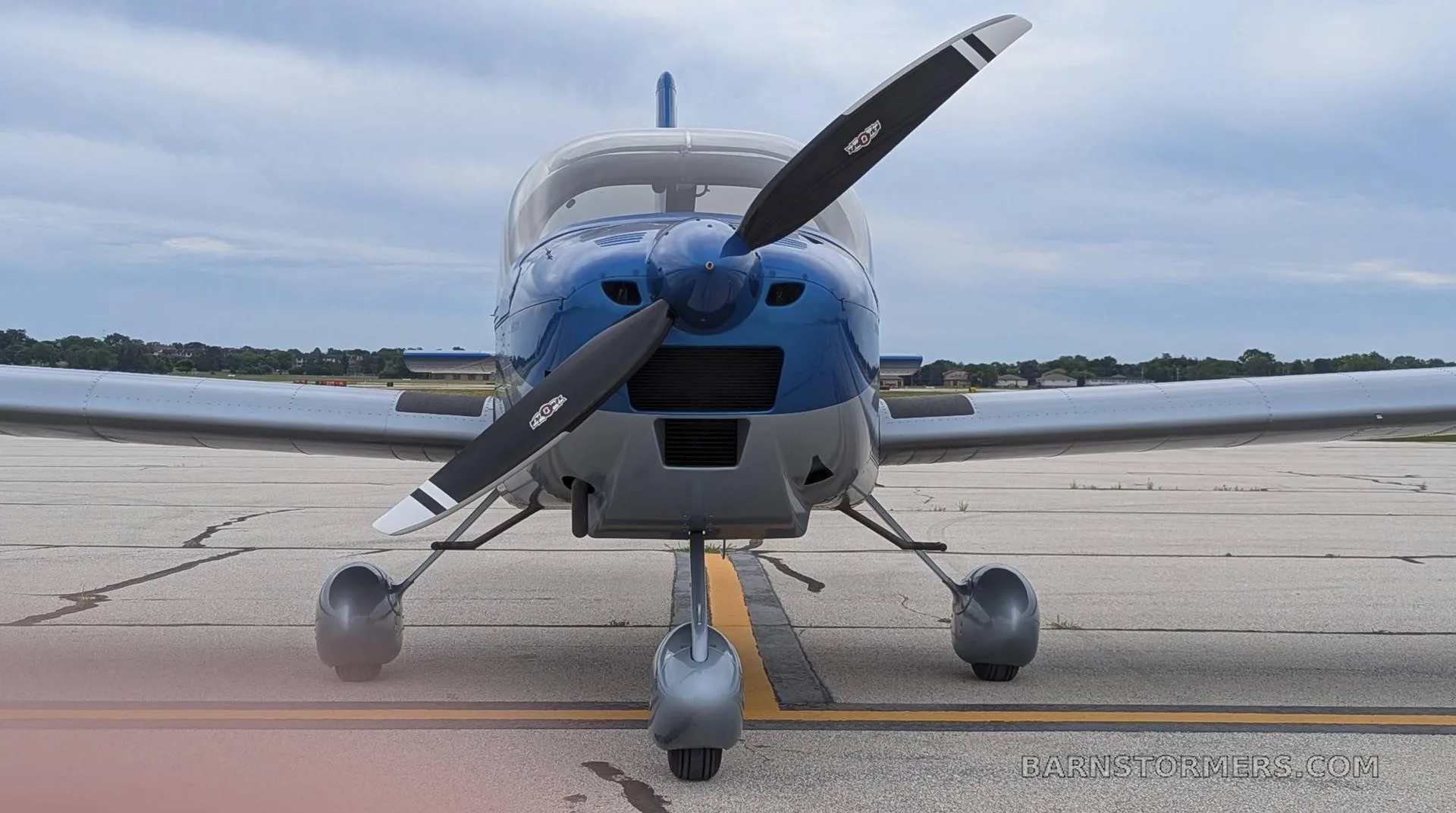 A blue and silver single-engine propeller aircraft, possibly a Van's RV, seen from the front on an airfield.