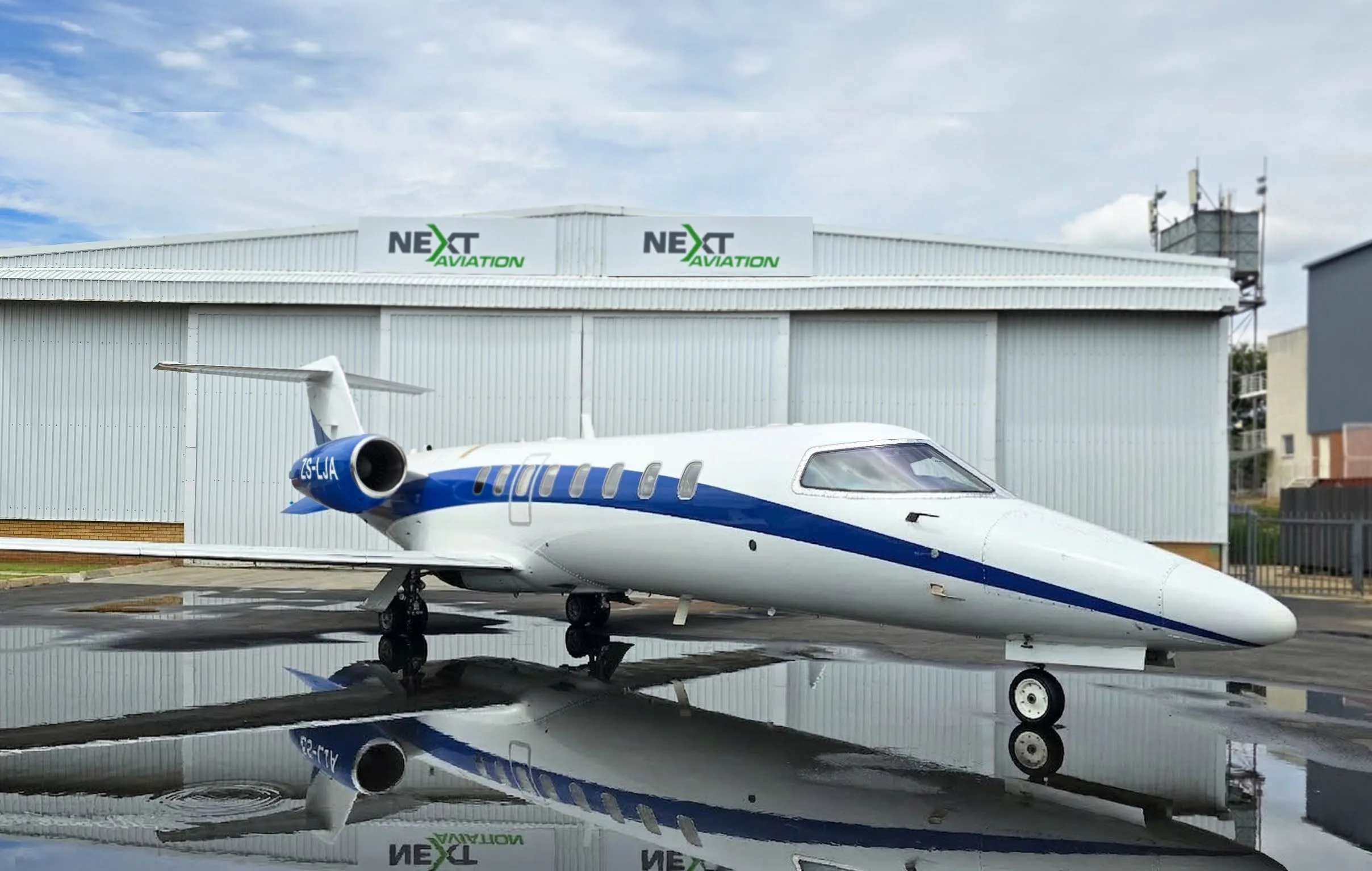 A white and blue Learjet, registration ZS-LJA, is parked on a wet tarmac in front of a hangar, reflecting in puddles.