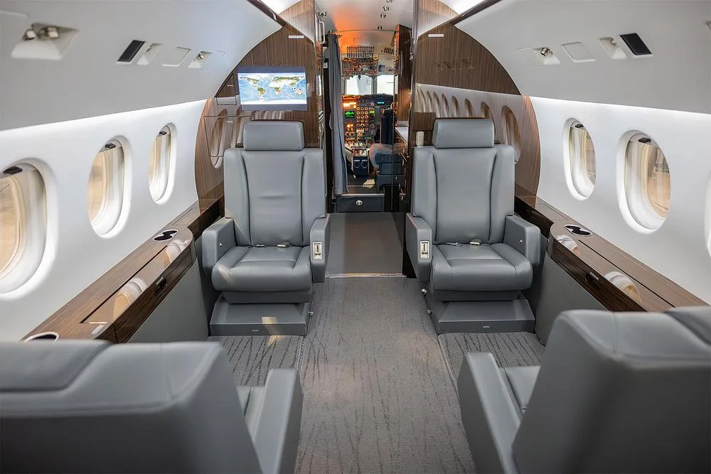 The luxurious cabin interior of a business jet, featuring grey leather seats, wood accents, and a screen displaying a world map, with the cockpit visible in the background.